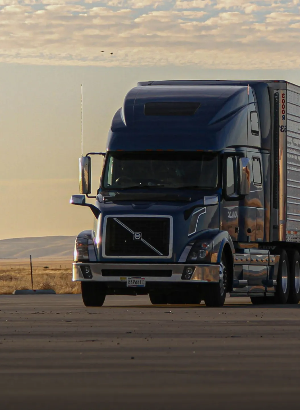 Blue Volvo semi truck parked on a flat paved area with hills and cloudy sky in the background.