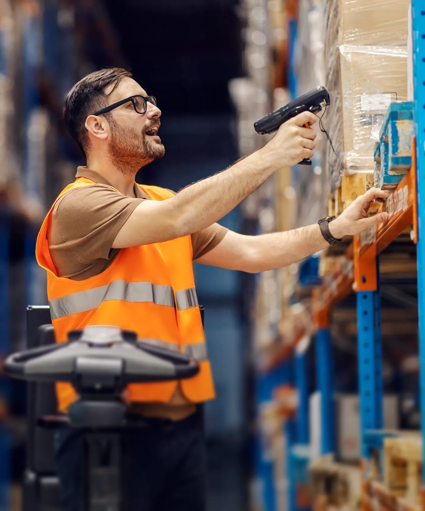 Warehouse worker in orange vest scanning a pallet on a high shelf with a handheld scanner.