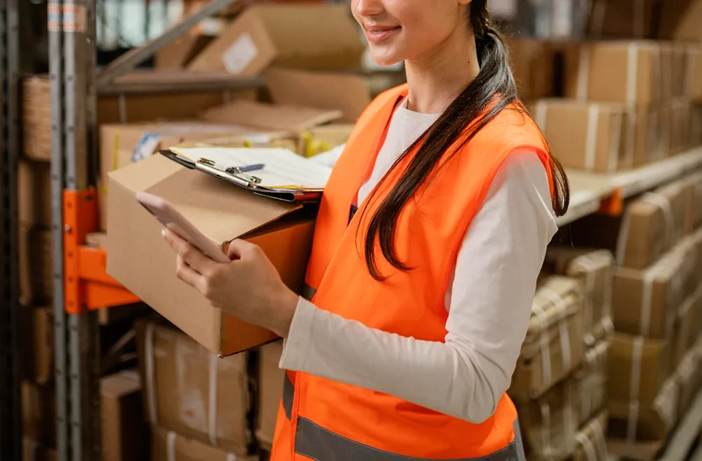 Warehouse worker in orange vest holding a box and smartphone with clipboard behind the box.