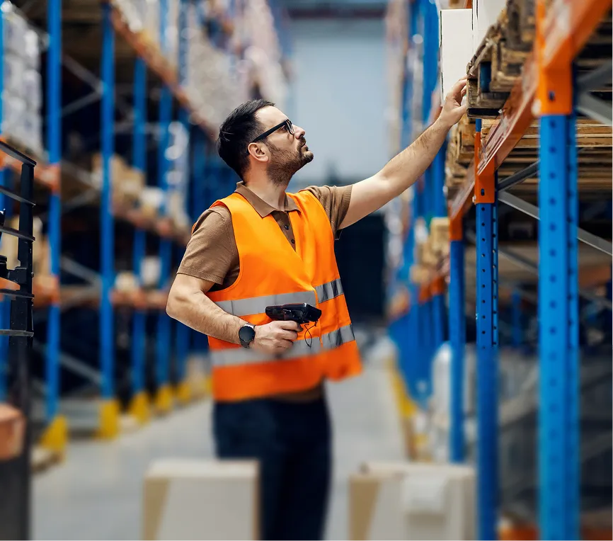 Warehouse worker in an orange safety vest scanning and checking items on high shelves.