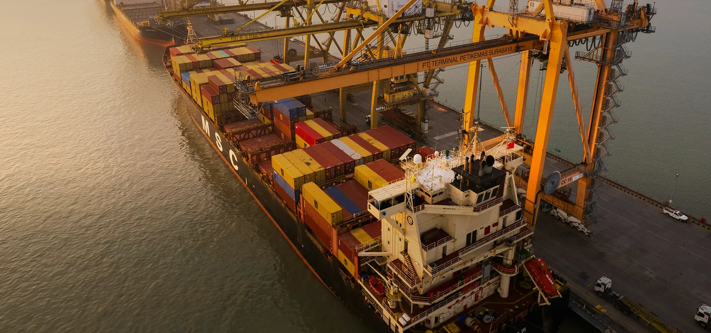 A container ship docked at a port with cranes loading and unloading cargo containers at sunset.